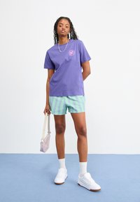 Camiseta de algodón morada con un logo circular, combinada con pantalones cortos a rayas verdes y azules. Zapatillas blancas y un bolso de color morado claro. Fondo plano.