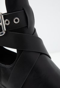 Zapato de cuero negro con correas cruzadas, que presenta una hebilla metálica y detalles de tachuelas. Textura suave y diseño de punta redonda.