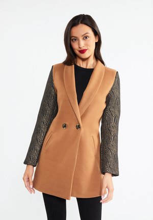 Femeie care poartă un palton fără mâneci de culoare cămilă, cu mâneci lungi negre texturate și pantaloni negri, stând pe un fundal alb simplu.