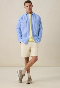 Lichtblauw overhemd met lange mouwen en twee borstzakken, gele t-shirt eronder, witte shorts en beige sneakers met grijze accenten.