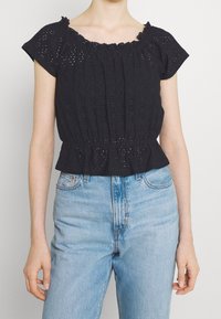 Svart topp med kort ärm, i ögonvitt tyg, med volangkrage och rynkad midja. Bärs med ljusblå jeans med hög midja.