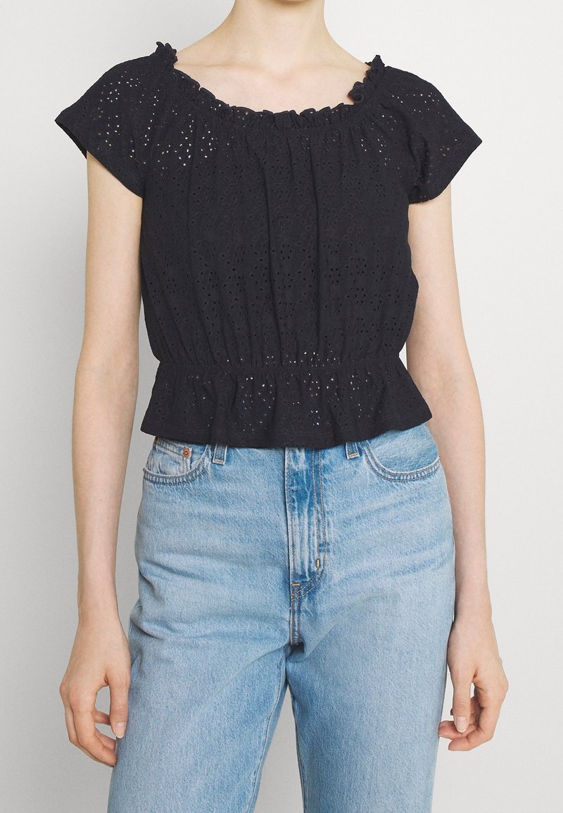 Svart topp med kort ärm, i ögonvitt tyg, med volangkrage och rynkad midja. Bärs med ljusblå jeans med hög midja.