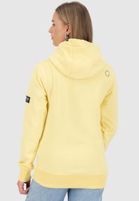 Vrouw met lang blond haar, gekleed in een lichtgele hoodie en lichtblauwe spijkerbroek, met haar rug naar de camera tegen een neutrale achtergrond.