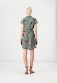 Abito floreale verde con maniche corte e vita elastica. Presenta motivi in rosa e verde scuro. La parte posteriore è leggermente più lunga di quella anteriore.