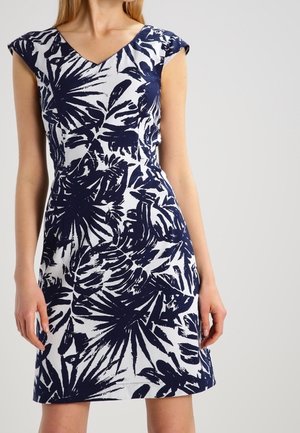 Mujer con un vestido blanco hasta la rodilla con un estampado de hojas tropicales azul marino y mangas cortas tipo capa.