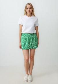 T-shirt bianco a maniche corte abbinato a una gonna verde con motivo floreale. Presenta una vita arricciata, lunghezza sopra il ginocchio e sneakers bianche.