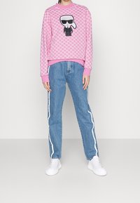 Roze sweatshirt met een cartoonfiguur die een zonnebril en een zwarte outfit draagt, gecombineerd met lichtblauwe jeans met witte zijkstrepen en witte sneakers.