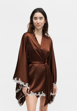 Γυναίκα που φοράει καφέ σατέν ρόμπα με φαρδιά μανίκια και λευκή δαντέλα στο τελείωμα, δεμένη στη μέση, στέκεται μπροστά σε απλό φωτεινό φόντο.