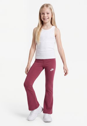 Unge pige smilende iført en hvid tanktop, bordeaux Nike bukser og hvide sneakers, stående mod en ensartet lys baggrund.
