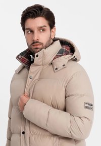 Beige puffermantel med avtagbar huva i rutigt mönster, tryckknappar och logotyp i ärmen. Slät textur och kviltat mönster.