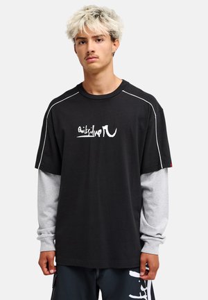 Junger Mann mit hellblonden Haaren trägt geschichtete schwarze und graue Langarmshirts sowie dunkle Hose mit weißem Grafikprint und steht vor weißem Hintergrund.
