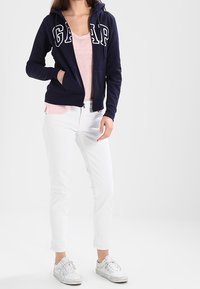 Sweat à capuche zippé bleu marine avec logo "GAP", porté sur un t-shirt rose clair. Associé à un pantalon blanc ajusté et des baskets blanches. Tissu lisse, design décontracté.