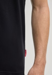 Zwarte katoenen t-shirt met een gladde textuur, voorzien van een zijlabel in het rood. Het ontwerp heeft een klassieke ronde hals en korte mouwen.