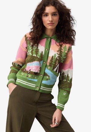 Kvinde med krøllet hår iført en knappet cardigan med design af skov, flod og lyserød van, kombineret med olivengrønne bukser.
