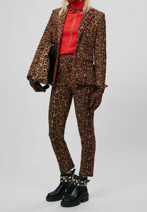 Kobieta w leopardowym żakiecie, spodniach, rękawiczkach, czerwonym golfie, trzymająca pasującą torbę w leopardowy wzór i czarne botki z ćwiekami.