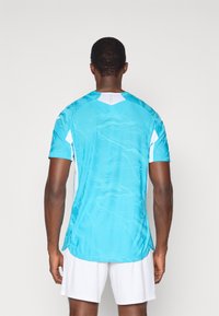 Μπλε αθλητική φανέλα με υφή, κοντά μανίκια και λευκές λεπτομέρειες. Η φανέλα έχει V-λαιμόκοψη και πλαϊνές σχισμές στο στρίφωμα.