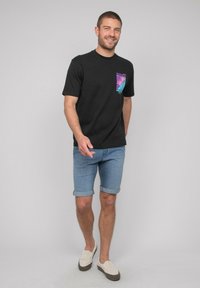 T-shirt noir avec un design coloré de palmier sur la poitrine, assortie à un short en denim bleu clair et des chaussures beiges à enfiler.