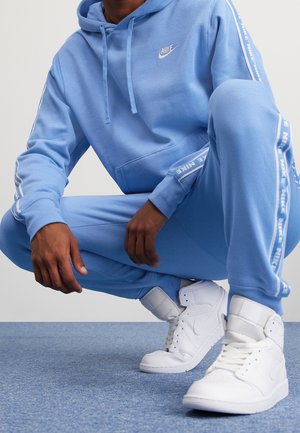 Osoba kucająca, ubrana w niebieską bluzę z kapturem Nike i dresowe spodnie z charakterystycznymi paskami oraz białe wysokie sneakersy Nike na niebieskim dywanie.
