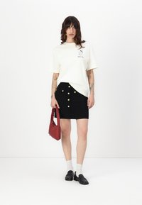 White graphic t-shirt paired with a fitted black skirt featuring gold buttons. Accessories include a red handbag and black loafers.
