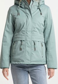 Vrouw in een lichtgroene hoodie met trekkoorden en ritszakken, gecombineerd met lichtblauwe jeans, tegen een neutrale achtergrond.