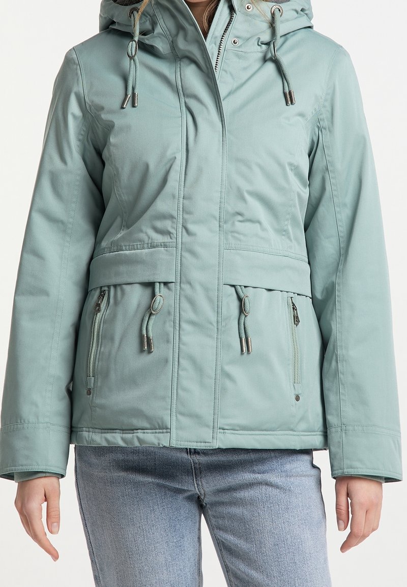 Vrouw in een lichtgroene hoodie met trekkoorden en ritszakken, gecombineerd met lichtblauwe jeans, tegen een neutrale achtergrond.