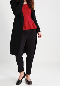Un long cardigan noir sur un top péplum rouge, associé à un pantalon noir et des chaussures plates noires. Le cardigan a des poches latérales.