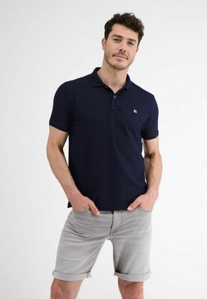 Mann trägt dunkelblaues Poloshirt und hellgraue, hochgekrempelte Shorts, steht mit den Händen in den Taschen vor einem schlichten weißen Hintergrund.