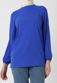 Maglia blu a maniche lunghe con scollatura a girocollo e polsini arricciati, realizzata in tessuto morbido. Indossata con jeans in denim blu, presenta una vestibilità comoda.