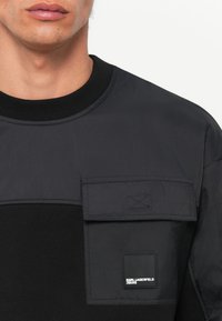 Hanorac negru cu un material texturat și un panou neted și lucios în partea de sus. Include o buzunar cu clapetă și detalii cu logo alb.
