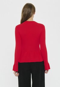 Maglione a coste rosso con orlo a peplum e maniche svasate. Texture liscia e design aderente. Pantaloni neri visibili nella parte inferiore.