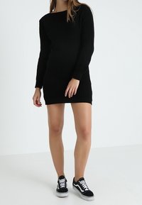 Robe tricotée noire à côtes avec des manches longues et un décolleté carré, associée à des baskets noires. Le tissu semble texturé et moulant.