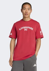 Muž v červenom tričku Liverpool FC a sivých teplákoch Adidas, stojí so jednou rukou v vrecku proti jednoduchému pozadiu.