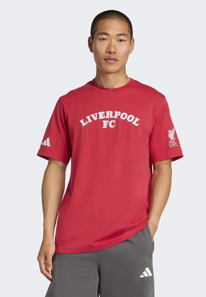 Muž v červenom tričku Liverpool FC a sivých teplákoch Adidas, stojí so jednou rukou v vrecku proti jednoduchému pozadiu.