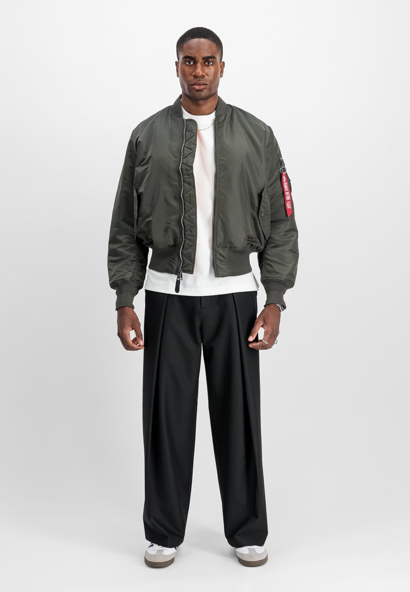 Chaqueta bomber de nylon verde oscuro con puños elásticos, cierre frontal de cremallera y bolsillos laterales. Combinada con pantalones anchos negros y zapatillas blancas.