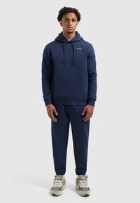 Navyblauwe hoodie met trekkoorden en logo, gecombineerd met bijpassende joggingbroeken. Het materiaal lijkt zacht met een gladde textuur. Draag met grijs gevoerde sneakers.