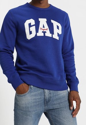 Osoba nosząca niebieską bluzę z białym i czerwonym logo "GAP Original", w połączeniu z jasnoniebieskimi jeansami.
