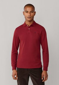 Polo à manches longues en coton rouge profond avec col et patte à trois boutons. Présente un petit logo bleu marine sur la poitrine.