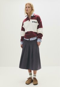 Donna bionda che indossa una felpa bordeaux e bianca di New York City sopra una maglietta a righe blu, una gonna midi plissettata blu scuro, calzini a righe e scarpe marroni.