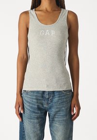 Top de tirantes de canalé gris claro con escote redondeado, que presenta el logotipo "GAP" en blanco. Combinado con jeans de mezclilla azul desgastados.