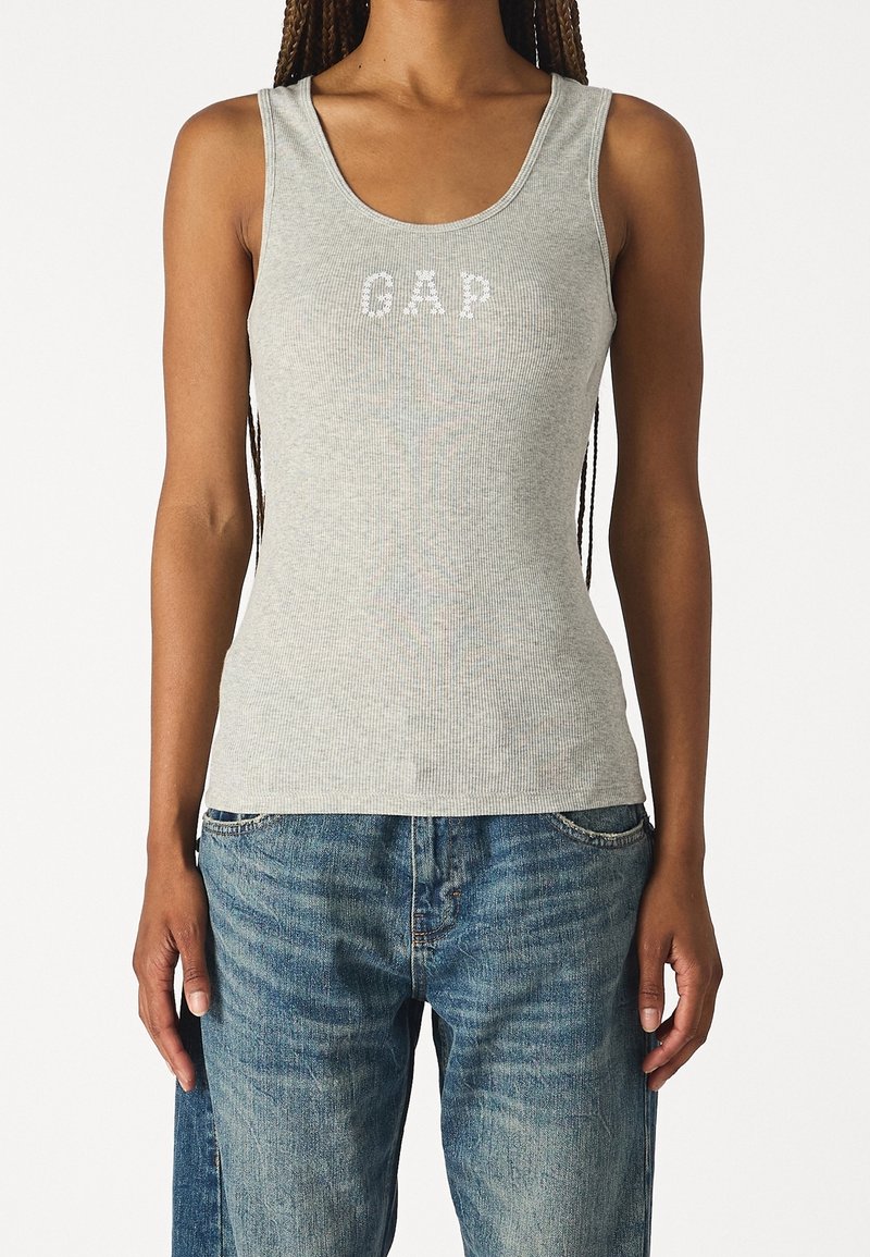 Top de tirantes de canalé gris claro con escote redondeado, que presenta el logotipo "GAP" en blanco. Combinado con jeans de mezclilla azul desgastados.