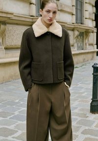 Veste en laine marron avec un col en peau de mouton, fermeture à boutons à l'avant et deux poches avant. Associée à un pantalon marron à jambes larges.