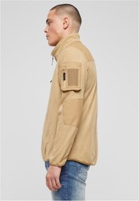 Világosbarna polárdzseki magas nyakkal, elöl cipzárral, texturált anyagból, és foltzseb a ujján. Megerősített vállrészekkel rendelkezik.