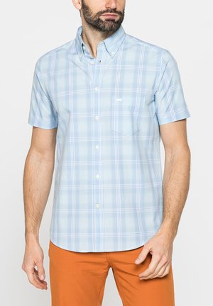 Uomo che indossa una camicia azzurro chiaro a maniche corte con motivo a quadri bianchi e tasca, abbinata a pantaloni arancioni.