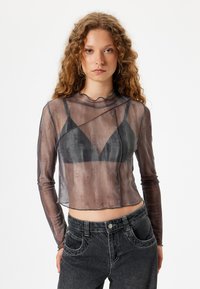 Haut transparent noir à manches longues, col festonné et design superposé sur un soutien-gorge noir. Associé à un jean foncé taille haute.