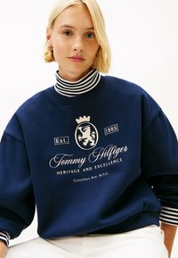 Granatowy sweter z białym wyszywanym logo przedstawiającym lwa i tekst. Na szyi widoczna warstwa z kołnierzykiem w paski. Materiał bawełniany.