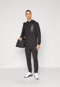 Svart hettegenser med grønn grafisk tekst, kombinert med matchende svarte joggebukser og hvite sneakers. Svart sportsbag holdes i venstre hånd.