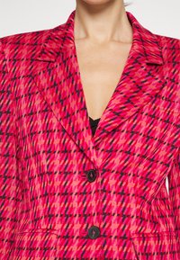 Femme portant un blazer rouge et rose à motifs avec des boutons noirs, laissant entrevoir un top noir en dessous, visible du cou à la taille.