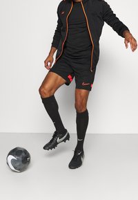 Svart idrottsoutfit med en kortärmad topp, shorts med röda detaljer, knähöga svarta strumpor och svarta fotbollsskor, tillsammans med en fotboll.
