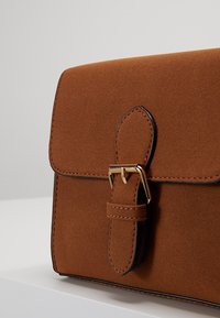 Brązowa zamszowa torebka o wyraźnie zarysowanym kształcie, wyposażona w klapkę zapięcia i złote akcenty oraz ozdobiona kontrastującymi przeszyciami.