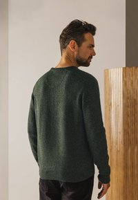 Donkergroene gebreide trui met een gestructureerd oppervlak, ronde halslijn en geribbelde manchetten. Gemonteerd met zwarte broek. Eenvoudig, klassiek ontwerp.